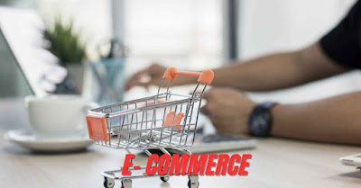A miniature shopping cart on a desk with a person working on a laptop and calculator. The image has the word "E-COMMERCE" on it, representing an online business for women from home.