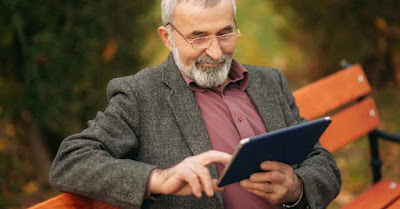 A retired man using a digital tablet while sitting on a park bench.